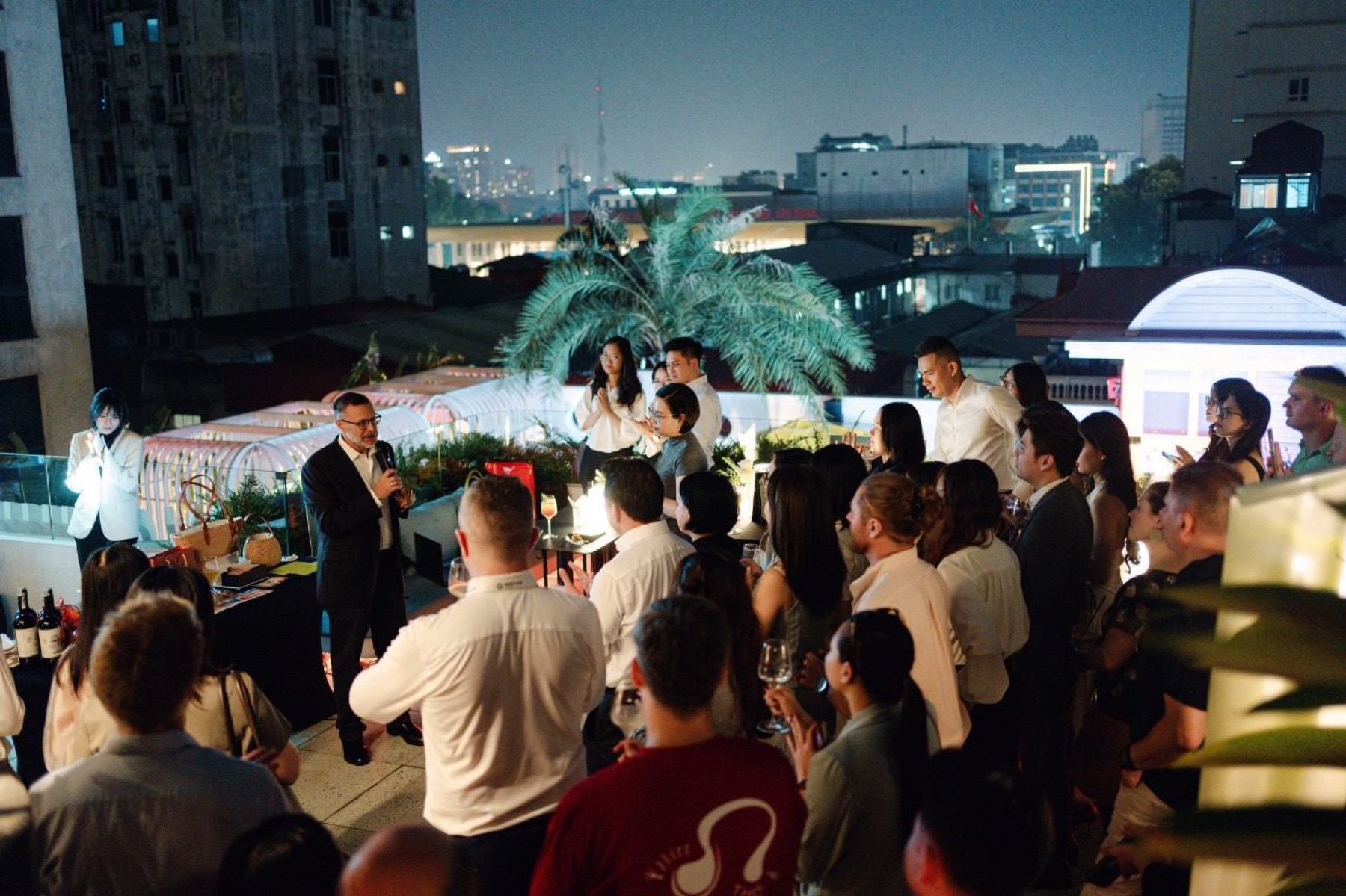 An evening rooftop reception in progress, a speaker addressing the room with the city behind